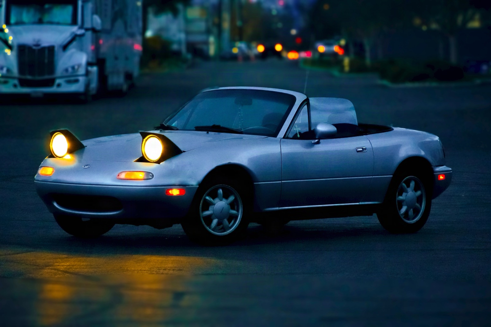 A 1991 Mazda Miata parked on a city street at night with a semi truck behind it